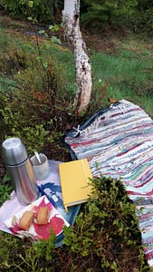 Koivun juurella räsymatto, pullakahveet ja keltainen muistikirja.